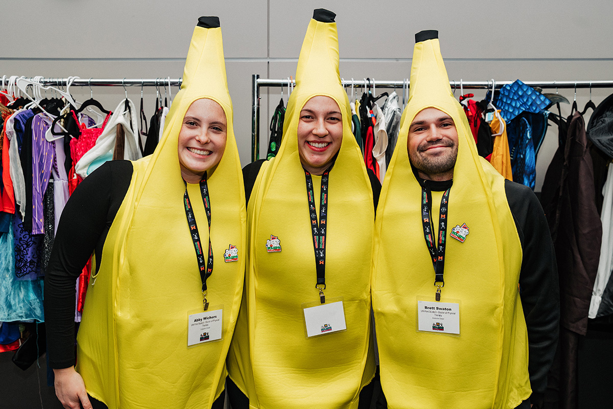 Three people wearing banana costumes stand in front of a rack of colorful clothing.