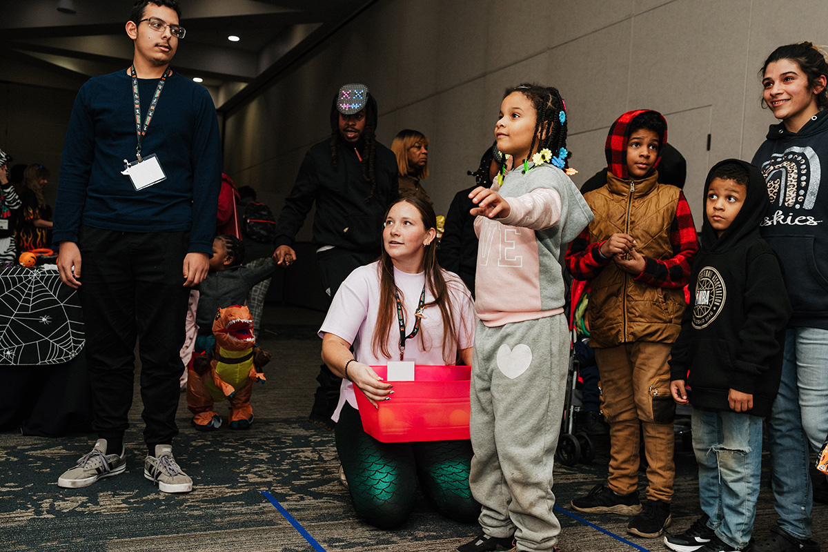 A group of children and adults in a festive setting, with one child pointing while another holds a red container.