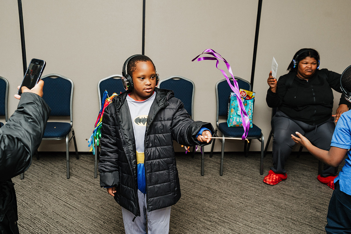 A child wearing a Batman shirt and headphones is holding a stick with colorful ribbons, while others are seated nearby.