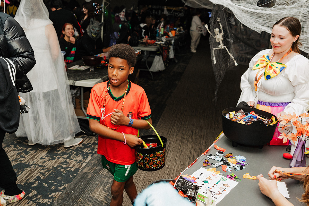 A child in an orange soccer jersey holds a black pumpkin-themed bucket while standing near a table with Halloween treats.