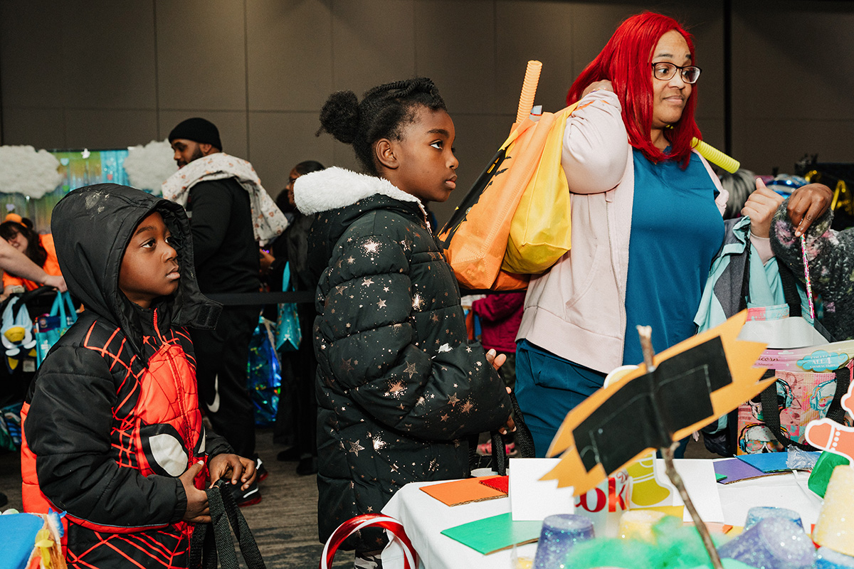 Children in costumes are interacting at a festive event, with colorful decorations and craft materials on a table.