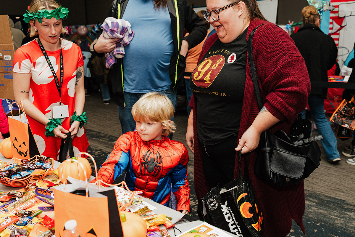 A child in a Spider-Man costume sits at a table decorated with pumpkins and candy, surrounded by adults in costumes.