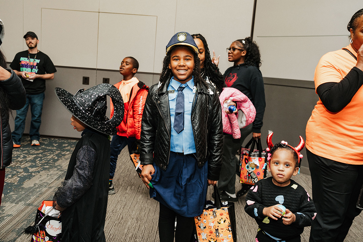 A group of children in costumes, including a girl dressed as a police officer and another wearing a witch hat, holding treat bags.