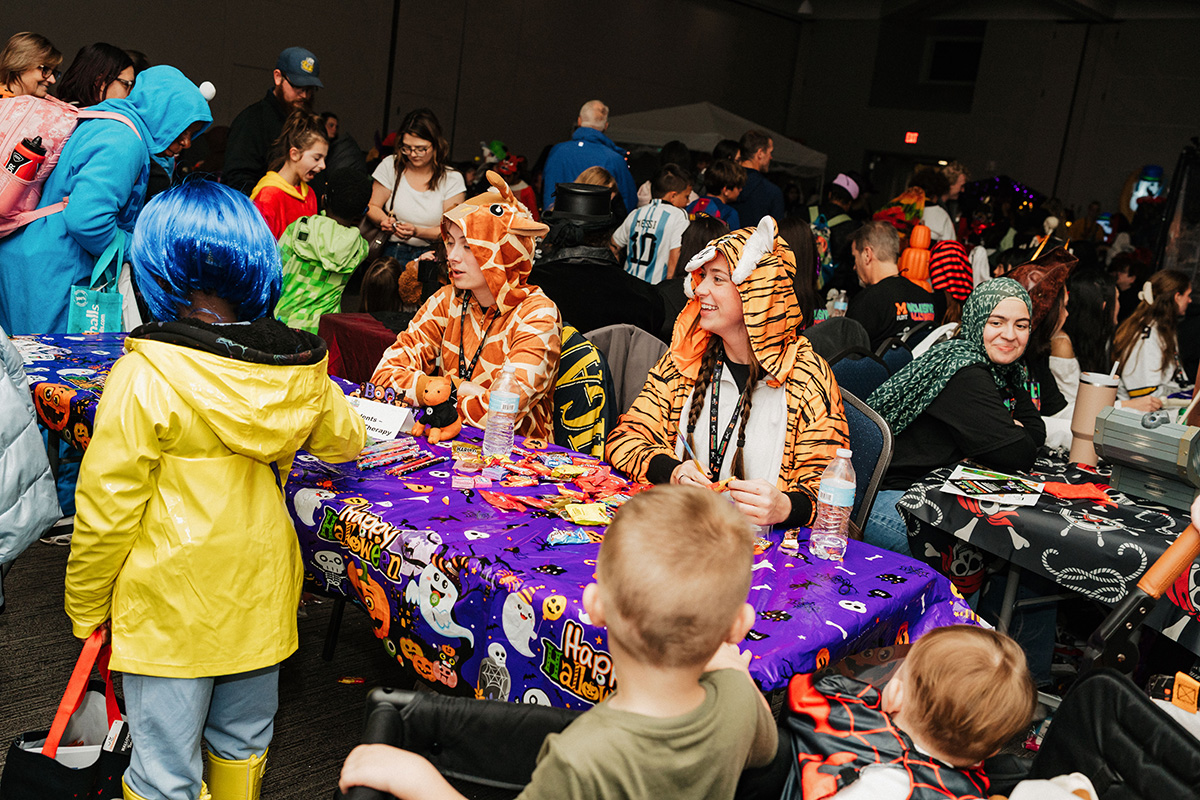 A crowded event scene with children in costumes, including two adults in animal onesies at a decorated table.