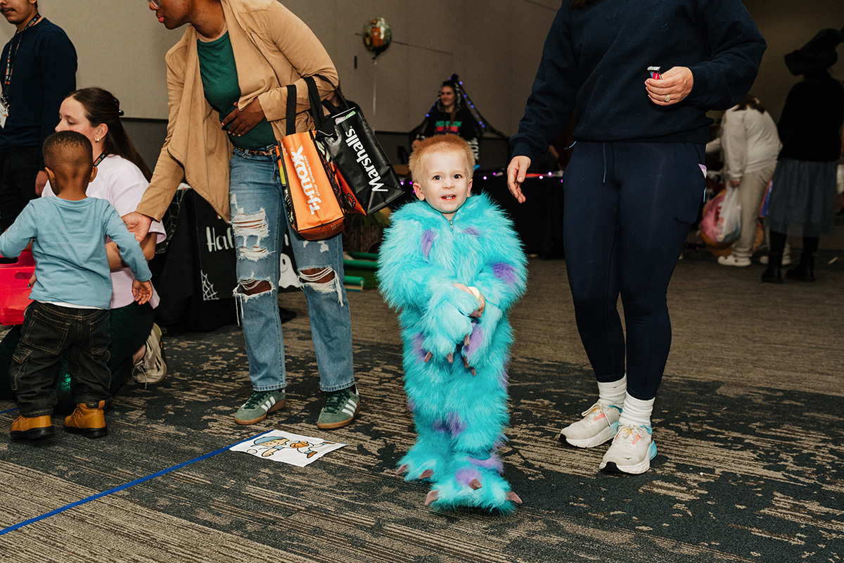 A child in a fluffy blue and purple costume stands among adults at a festive event, with decorations in the background.