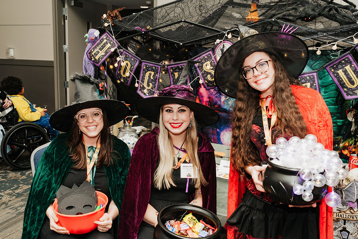Three individuals dressed as witches, holding bowls of candy, in front of a Halloween-themed backdrop with decorations.
