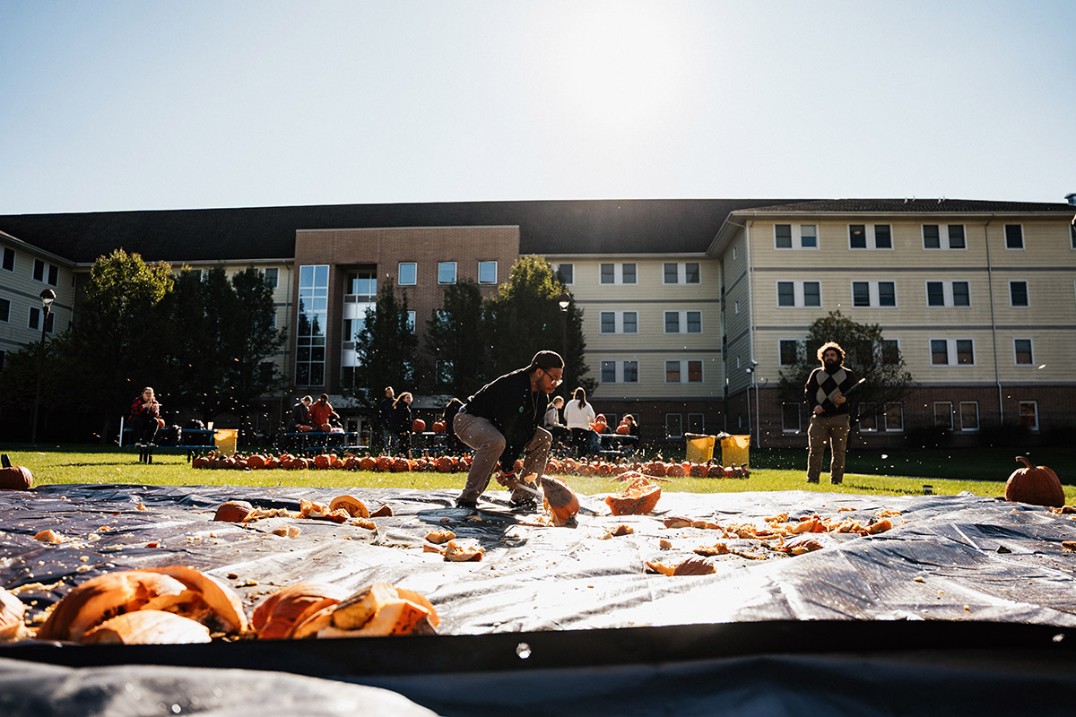 A person is carving a pumpkin on a tarp, with scattered pumpkin pieces around, and a group of people and pumpkins in the background.