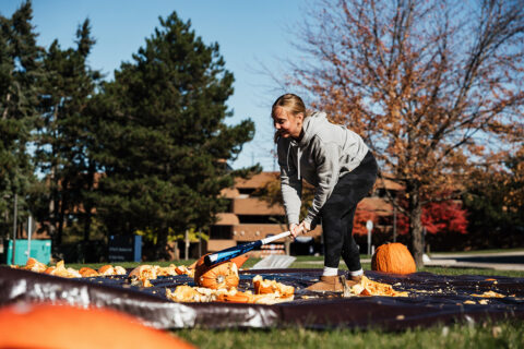 A person in a gray hoodie is smashing pumpkins on a black tarp in a park, surrounded by trees and autumn foliage.