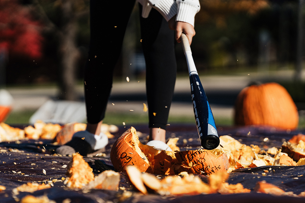 A person in black leggings and slippers swings a baseball bat at smashed pumpkins, with pumpkin pieces and seeds flying.