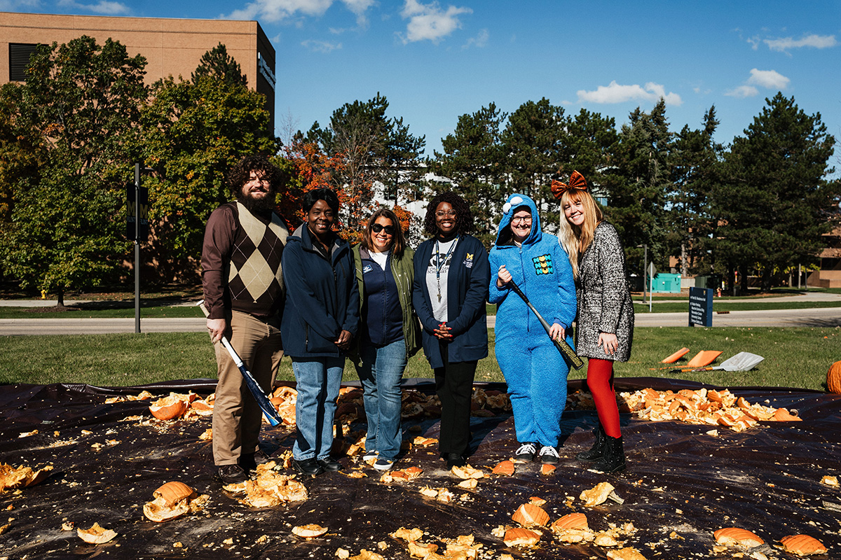 A group of six people standing on a ground covered with pumpkin remnants, surrounded by trees and a building in the background.