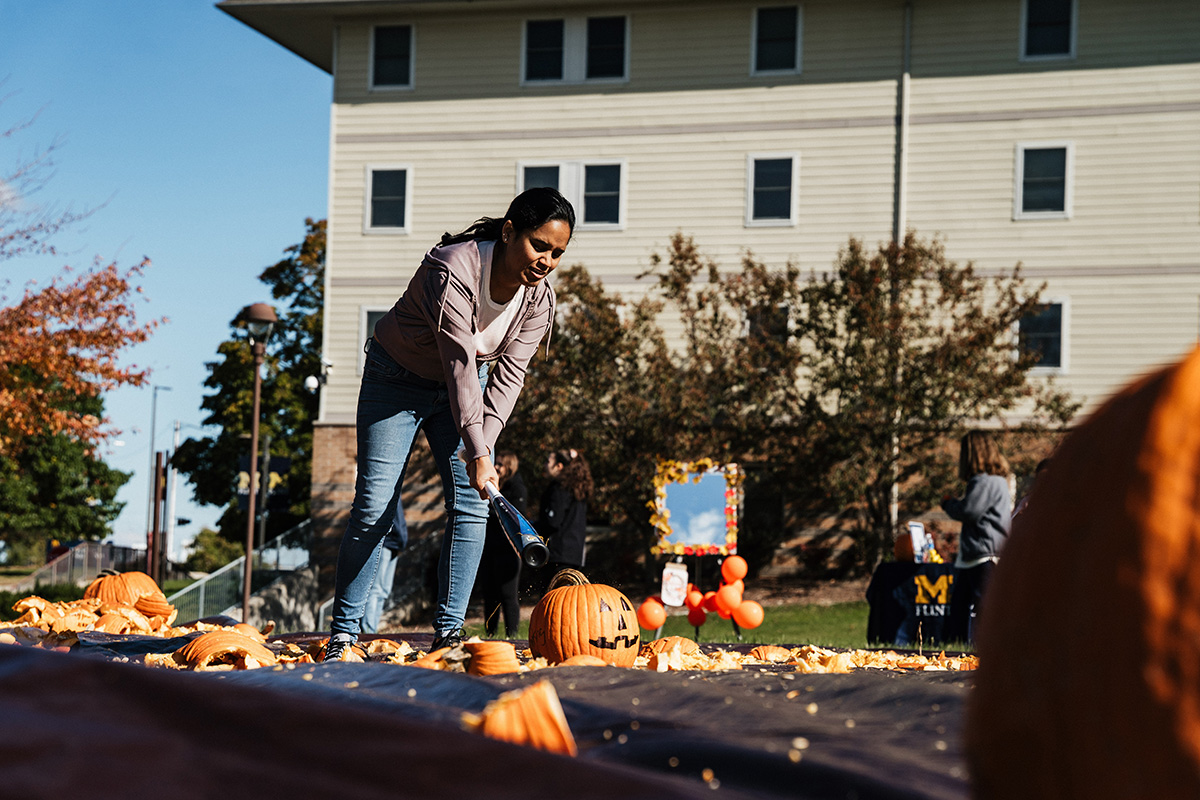 A person is using a tool to carve pumpkins on a table covered with pumpkin scraps, with a building and people in the background.