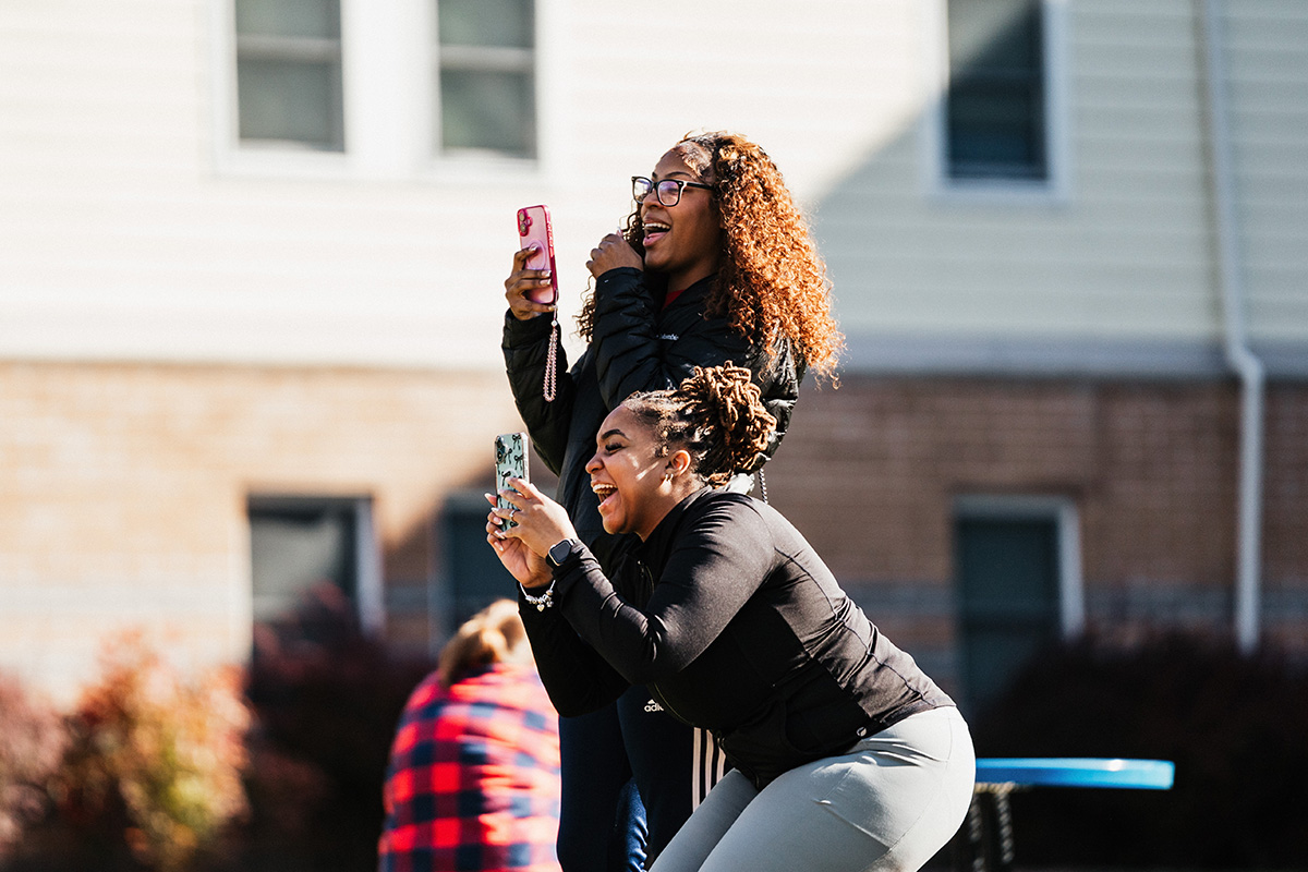 Two women taking selfies outdoors, one standing and the other squatting, with a blurred background.