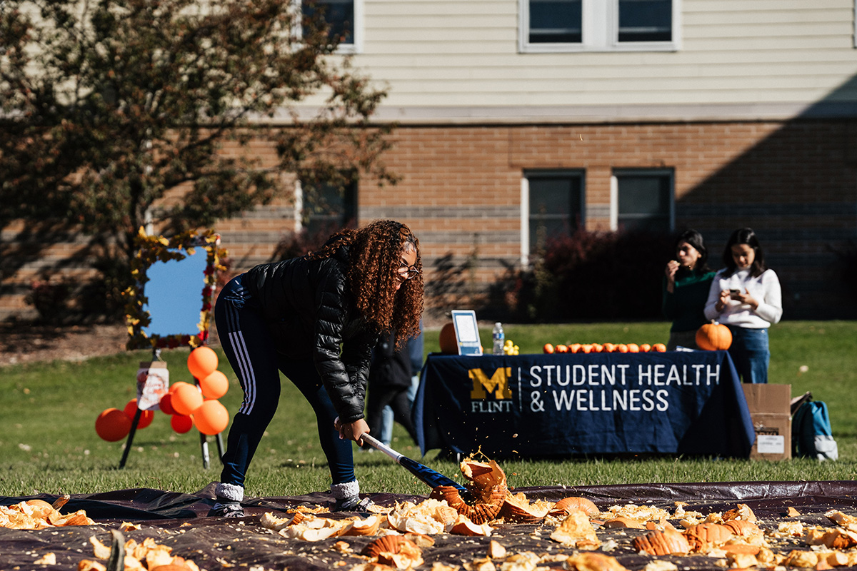 A person shoveling pumpkin guts at an outdoor event with a "Student Health & Wellness" table in the background.