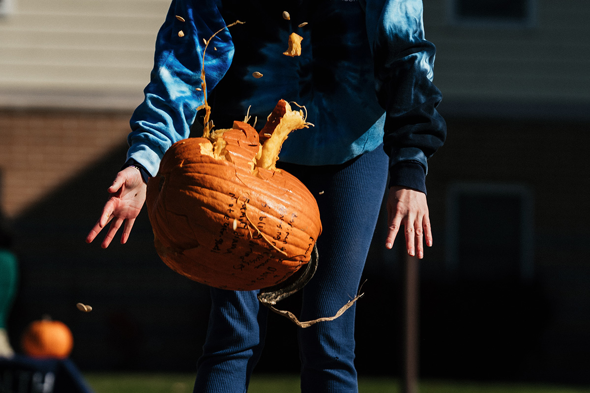 A person in a blue tie-dye sweatshirt is dropping a pumpkin, with pumpkin guts and seeds flying out.