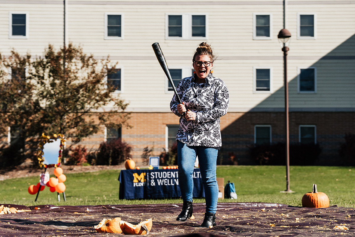 A person stands on a tarp in a field, holding a baseball bat, ready to hit a pumpkin. In the background, a table displays a sign for "Student Health & Wellness."