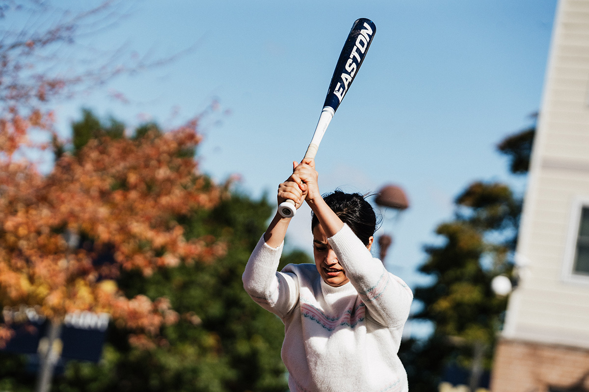 A person in a white sweater is preparing to swing a baseball bat outdoors, with trees and buildings in the background.