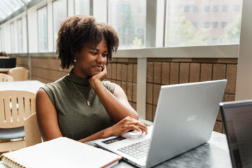 A person sitting at a table using a laptop, with a notebook and pen nearby, in a well-lit indoor space.