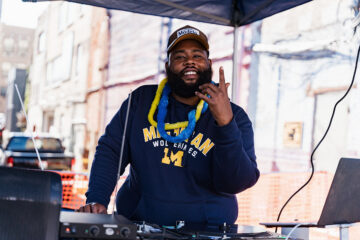 A DJ wearing a Michigan Wolverines hoodie and a colorful lei gestures while performing at an outdoor event.
