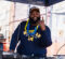 A DJ wearing a Michigan Wolverines hoodie and a colorful lei gestures while performing at an outdoor event.