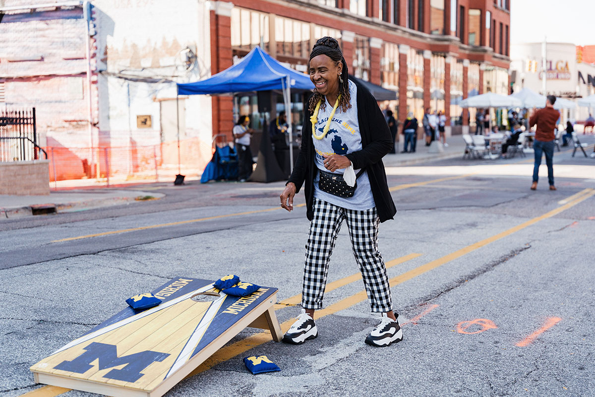 A person wearing a Michigan-themed outfit stands near a cornhole game set up on a street during an outdoor event.