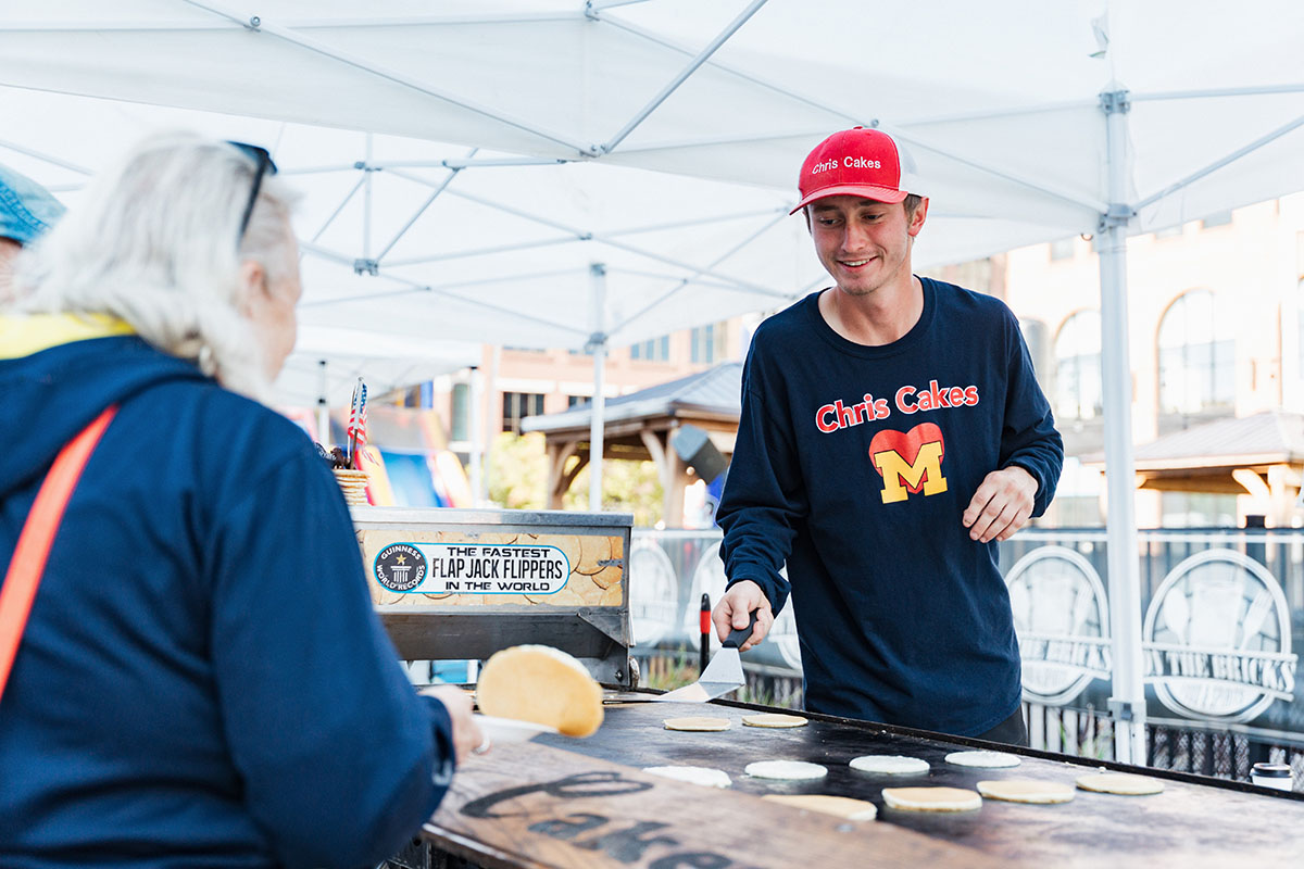 A person in a red hat and dark shirt with "Chris Cakes" is flipping pancakes at a food stall, with a sign that reads "THE FASTEST FLAP JACK FLIPPERS IN THE WORLD."