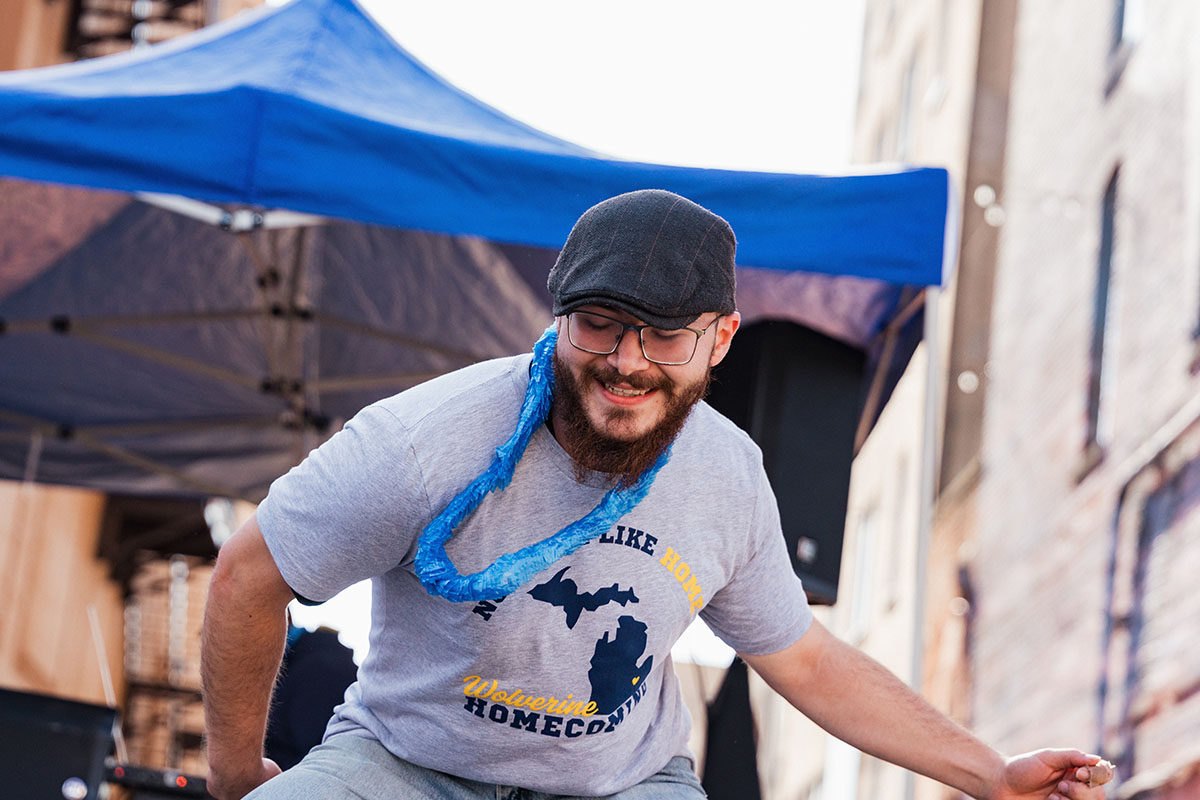 A person wearing a gray t-shirt with the text "Michigan Like Home Wolverine Homecoming" dances under a blue tent.