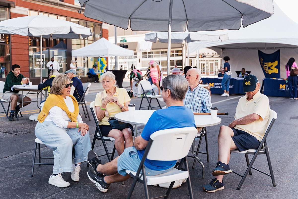 A group of older adults sitting around tables at an outdoor event, engaged in conversation. Tents and a banner are visible in the background.