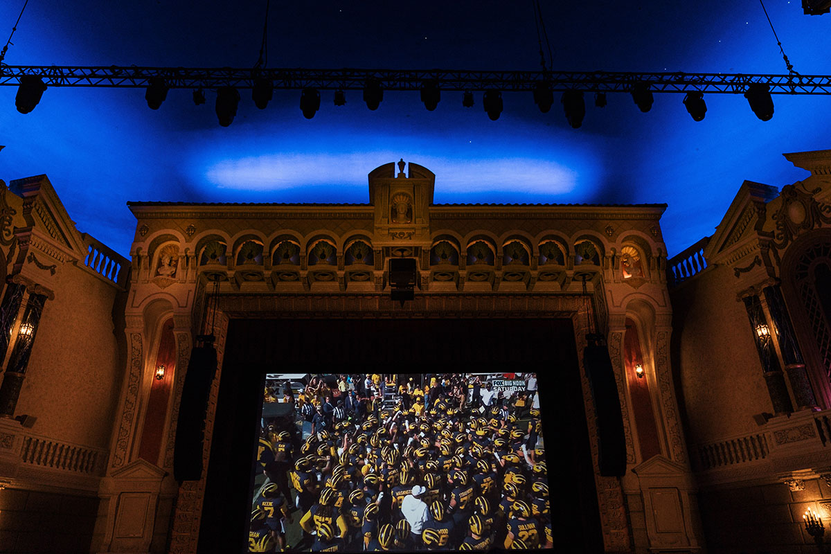 A large screen displays a crowd of people in yellow and black uniforms, set in an ornate theater with a blue ceiling.