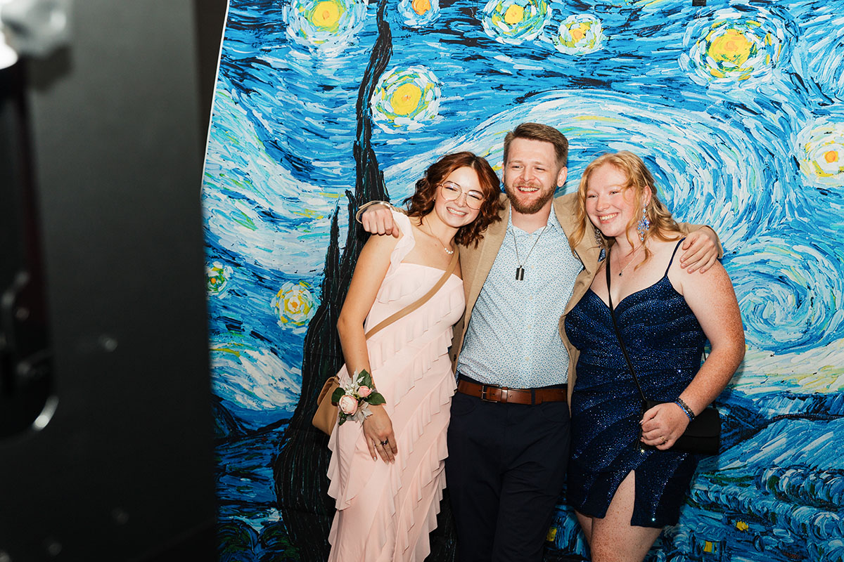 One male student in the middle flanked by two female students posing for a photo in front of the starry night backdrop.
