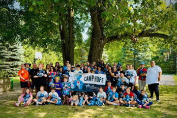 A large group of children and adults gathered outdoors under trees, holding blue teddy bears and a sign that reads "CAMP HOPE."