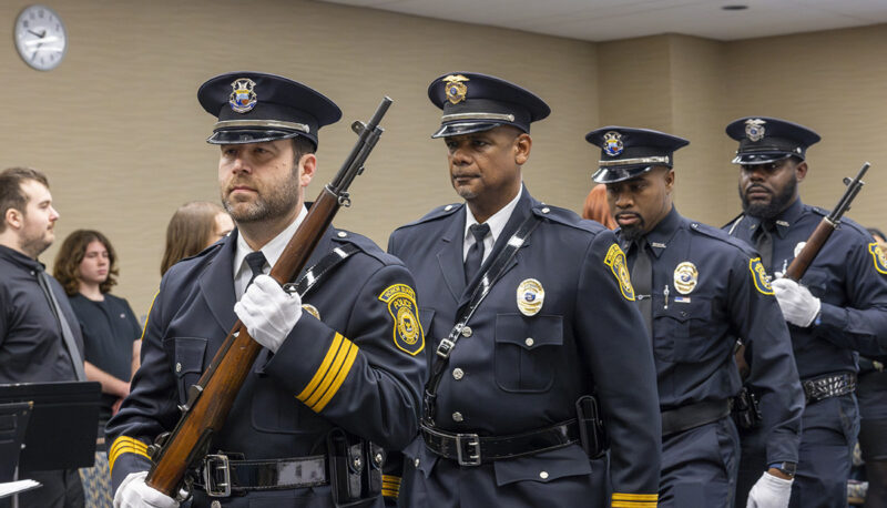 A group of police officers in uniform, some holding rifles, marching in a formal setting.