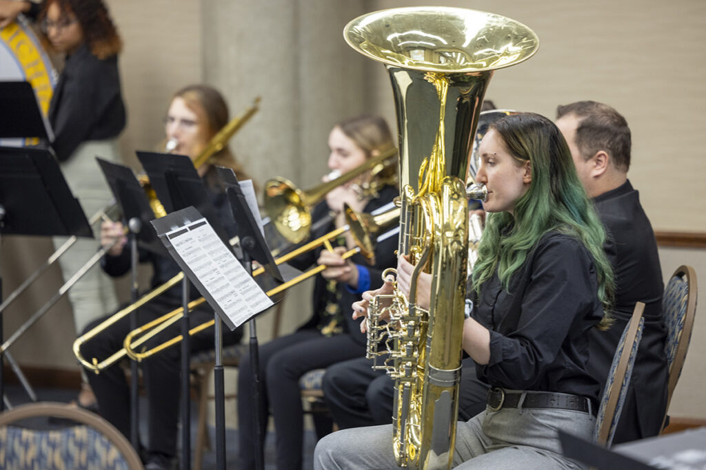 members of the UM-Flint Brass Ensemble wearing black dress clothes and playing their instruments. The photo focuses on a female student with green highlights playing the tuba. 