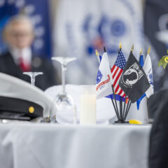 A table setting featuring military hats, small flags representing different branches of the military, a candle, and a rose with a yellow ribbon.