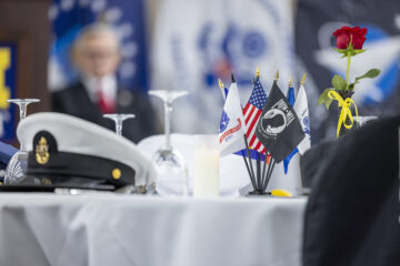 A table setting featuring military hats, small flags representing different branches of the military, a candle, and a rose with a yellow ribbon.