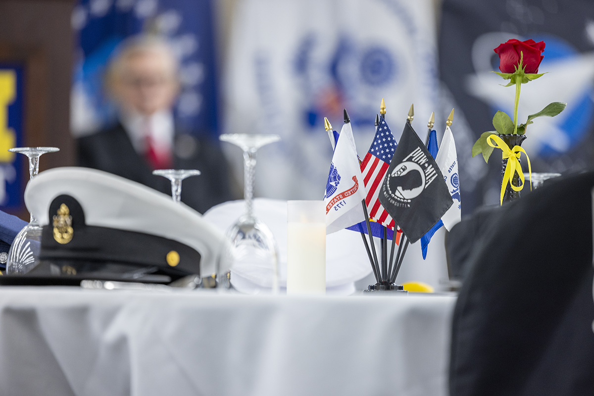A table setting featuring military hats, small flags representing different branches of the military, a candle, and a rose with a yellow ribbon.