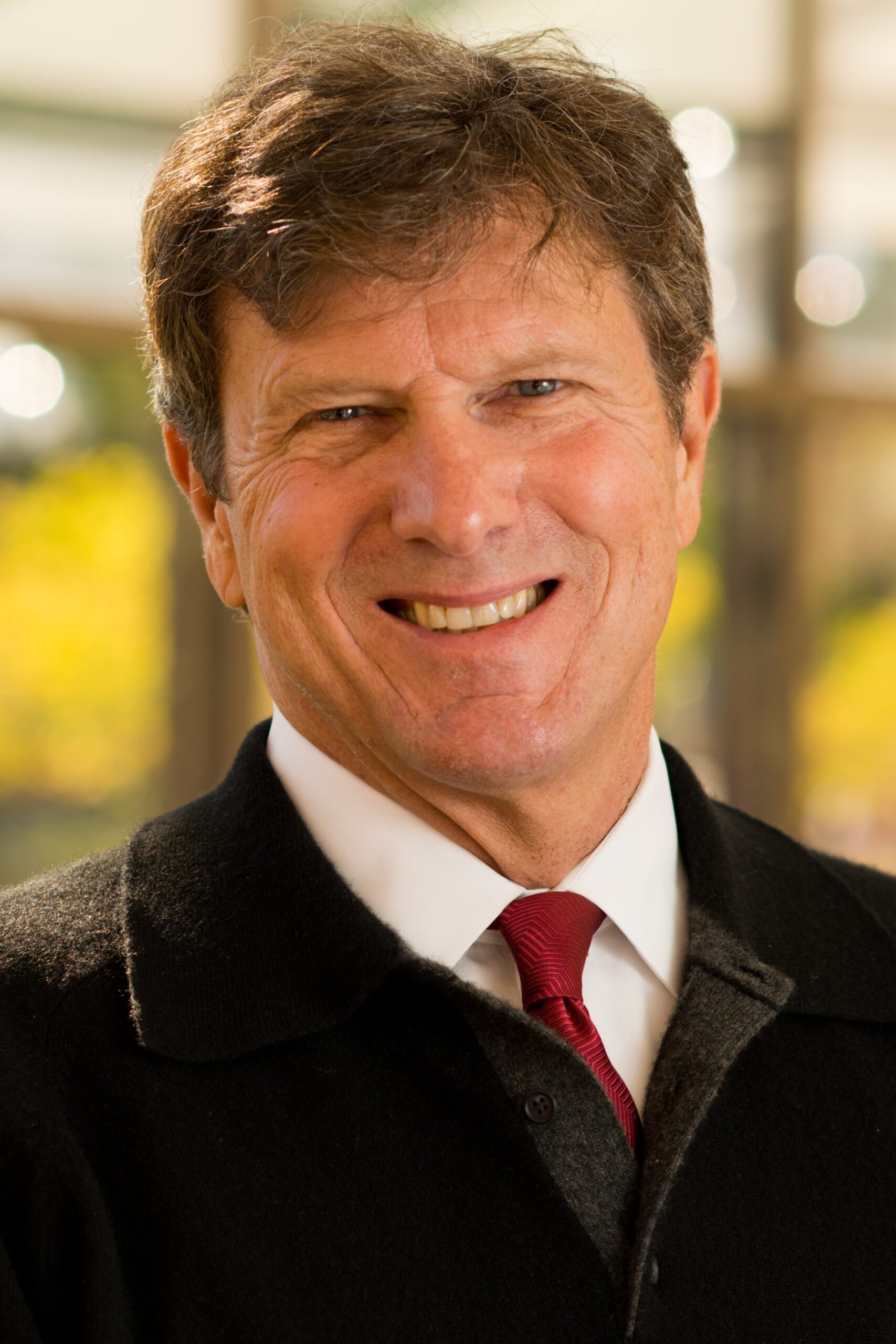 A man with short brown hair smiles for a portrait. He is wearing a black sweater overtop a white dress shirt and red tie.
