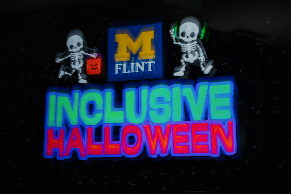 Colorful sign reading "INCLUSIVE HALLOWEEN" with skeletons, one holding a pumpkin bucket and the other wearing headphones.