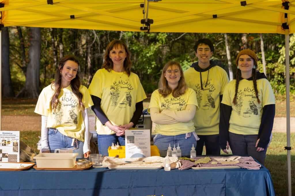 A group of five people wearing yellow "Bioblitz 2025" t-shirts stand behind a table with nature observation materials under a yellow canopy.