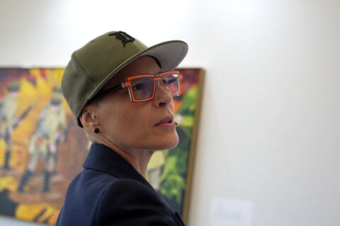A person wearing a green cap and glasses stands in front of a colorful artwork in an art gallery.