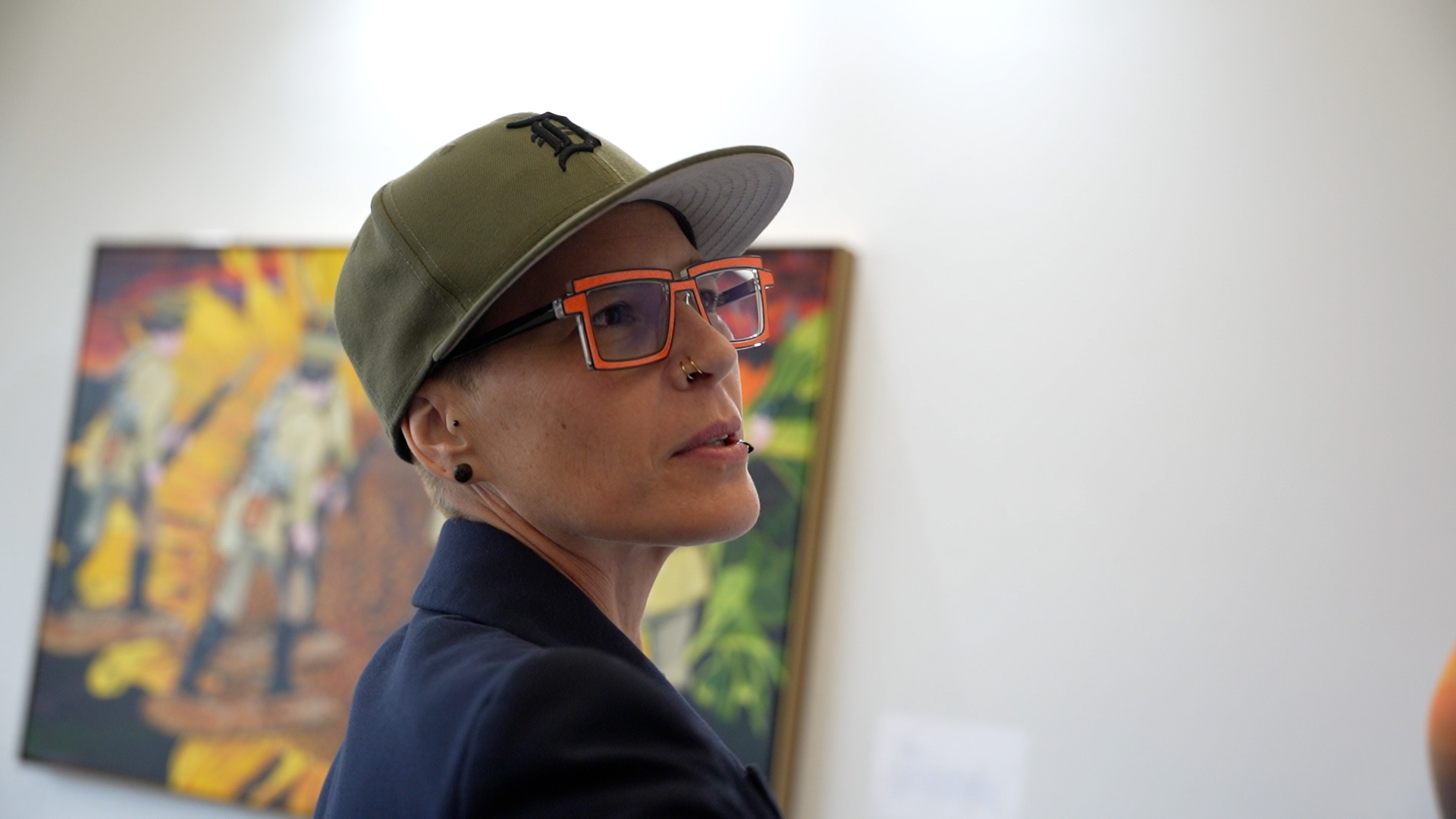 A person wearing a green cap and glasses stands in front of a colorful artwork in an art gallery.