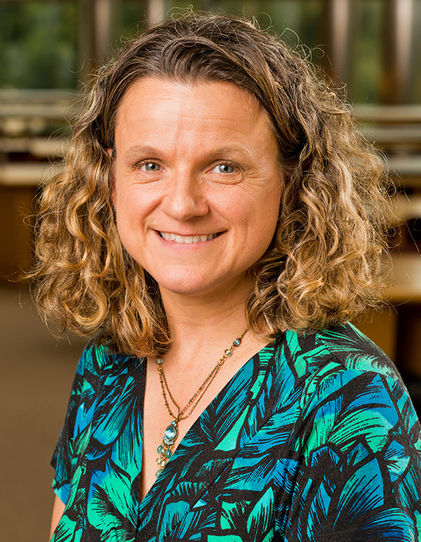 A person with curly hair wearing a green and blue patterned top and layered necklaces, standing indoors.