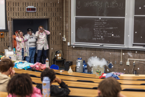 A chemistry demonstration in a classroom with students in lab coats reacting to a cloud of vapor and a chalkboard listing activities.