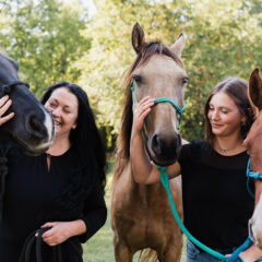 Two women are standing outdoors, each holding a horse. One horse is dark gray and the other is light brown with a white stripe.