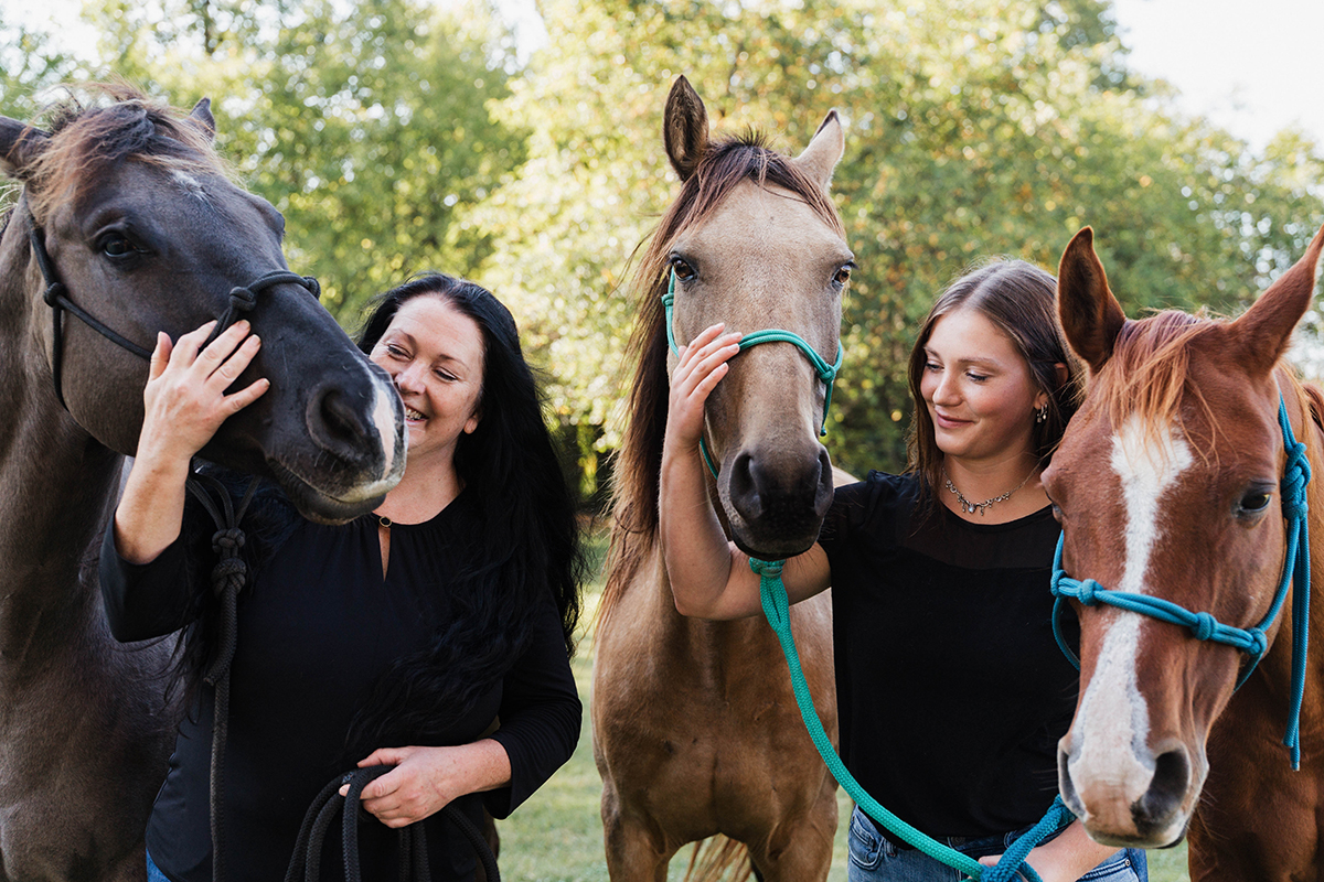 Two women are standing outdoors, each holding a horse. One horse is dark gray and the other is light brown with a white stripe.