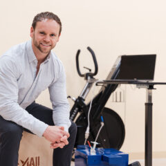 Curt Fennell sitting on a wooden box in a gym-like setting, with an exercise bike and a laptop nearby.