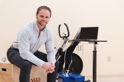 Curt Fennell sitting on a wooden box in a gym-like setting, with an exercise bike and a laptop nearby.