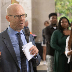 A speaker gestures while holding notes and addressing a group of people in a blurred background.