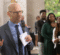 A speaker gestures while holding notes and addressing a group of people in a blurred background.