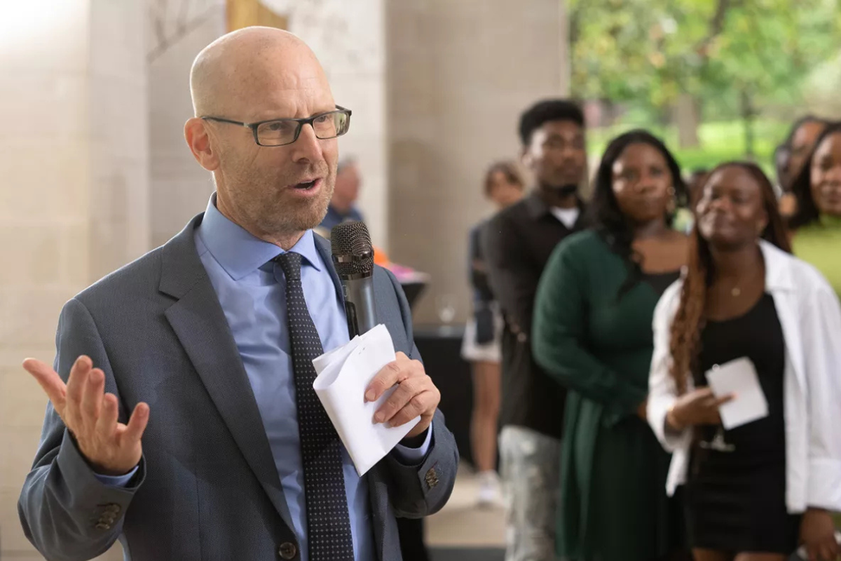 A speaker gestures while holding notes and addressing a group of people in a blurred background.