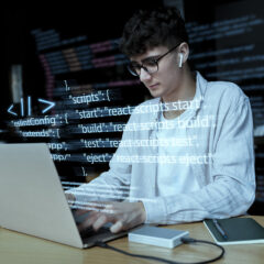 A person working on a laptop with code displayed on the screen, including commands for React scripts.